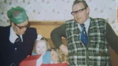 Fotografia colorida de uma reunião de família na década de 1970. Anthony Littler, um homem corpulento usando óculos, uma camisa xadrez verde sem mangas e uma gravata azul, aparece sentado e sorrindo em um sofá. À sua esquerda, um homem mais velho de óculos e chapéu de festa de papel verde se inclina em direção a uma menina pequena, de vestido azul, que abre um grande presente embrulhado em papel vermelho.