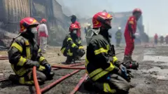 İran'ın Bender Abbas kentinde ülkenin en büyük ticari limanı olan Şehit Recai Limanı'ndaki yangına müdahale eden itfaiyeciler yere oturmuş durumda, hortum tutarak uzağa bakıyor. Fotoğraftaki herkes maskeli
