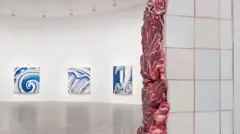 A foto mostra uma galeria de arte contemporânea de paredes brancas e espaço amplo. Em primeiro plano, uma coluna revestida de azulejos claros parece rasgada, revelando formas vermelhas e orgânicas que lembram carne ou vísceras, criando um contraste entre a superfície limpa e o interior brutal da estrutura. Ao fundo, três quadros com pinceladas azuis inspiradas em azulejos portugueses estão expostos na parede curva do salão iluminado.