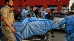 На каталке лежит человек под синим покрывалом, его везет медик в синем халате, рядом несколько человек, некоторые в синей медицинской одежде, некоторые в военной форме