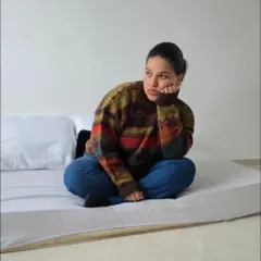 Rebeca Alvarez, uma mulher jovem vestindo calça jeans e malha colorida, sentada num colchão no chão, com uma expressão frustrada no rosto
