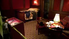Un niño observa destrás de una barrera una exposición en un museo de un camarote de barco antiguo, enchapado en madera, con una cama, una mesa redonda con una lámpara, libros y una taza de té y dos asientos a su alrededor