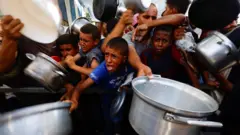 Espremidas ao centro, várias crianças palestinas tentam conseguir comida segurando grandes panelas de metal em um ponto de distribuição de alimentos no centro da Faixa de Gaza