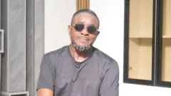 Nollywood actor, Emeka Ike siddon dey look inside camera, im wear gery colour shirt and dark eye glass.