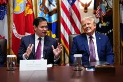 Rubio e Trump sentados diante de mesa, em frente a bandeiras; Rubio gesticula enquanto fala, e Trump sorri olhando para a frente
