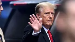 Donald Trump saludo de lado. Viste traje oscuro y corbata roja.