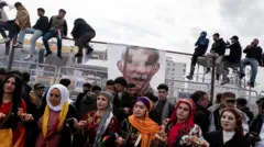 Kadınlar önde halay çekiyor, arkada genç erkekler. Bazıları tellerin üzerine oturmuş. Tellerde bir Abdullah Öcalan posteri asılı. 