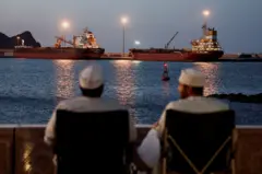 Dos buques cisterna permanecen anclados el 9 de marzo de 2026. En primer plano, dos hombres con túnicas blancas están sentados en sillas, contemplando el canal y los barcos.
