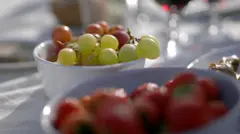 Tigela branca cheia de uvas brancas e tintas sobre uma mesa coberta com toalha branca. Outra tigela contém morangos vermelhos, levemente fora de foco. O fundo mostra utensílios de vidro borrados e outros produtos alimentícios, sugerindo uma refeição em ambiente externo com luz natural.