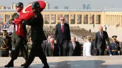 El presidente Recep Tayyip Erdogan sube los escalones del Mausoleo de Atatürk en Ankara durante una ceremonia del Día de la Victoria el 30 de agosto de 2016, flanqueado por funcionarios del gobierno mientras soldados portan una corona de flores con la forma de la bandera turca.