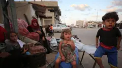 Gazze'de bir yol kenarında ikisi kız biri erkek üç küçük çocuk evlerinden kalan eşyalara oturmuşlar. Arkada bir yetişkin başörtülü kadın var