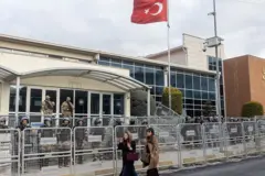Mahkeme salonu önünde demir parmaklıklar ve polis. İki kadın önde yürüyor. Yukarıda Türkiye bayrağı var.