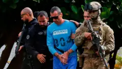 Marcola, líder do PCC, vestido com uma roupa azul de interno do presídio, escoltado por forças de segurança.