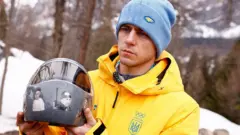O atleta de skeleton ucraniano Vladyslav Heraskevych segura seu capacete, que mostra vítimas da guerra do seu país contra a Rússia, nos Jogos Olímpicos de Inverno de 2026