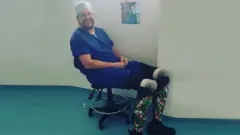 Nil Hoper sedi na stolici u plavoj uniformi i sa naočarima, sa protetskim udovima. Protetski udovi imaju šaru velškog zmaja i bele su, crvene i zelene boje. On se osmehuje u kameru.