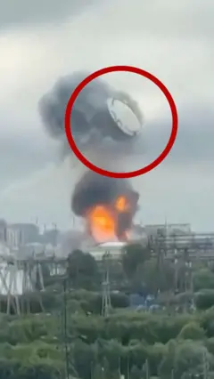 තෙල් ටැංකියක් ගිනි ගන්න අතර එහි පියනක් වීසි වෙන ආකාරය රතු පාටින් රවුම් කර තිබේ.