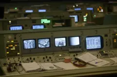 Sala de controle de voo da missão Apollo, em Houston, aberta à visitação pública, com monitores ligados, fichários, folhas de anotações, uma caneca e um cinzeiro com cigarros dentro