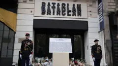 「BATACLAN（バタクラン）」という看板が据えられたホールの玄関前に、男女の衛兵が立っている。玄関前には事件の犠牲者の名前が書かれたボードが置かれている。玄関のガラスのドアには、献花の花束が山と積まれている