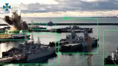 Военный порт с несколькими боевыми кораблями у причалов. На заднем плане – взрыв с большим столбом дыма и обломками возле одного из судов. На воде видны фрегаты и катера, а над горизонтом – облачное небо. На изображении есть зеленые прямоугольные рамки, которые выделяют корабли и объекты, что свидетельствует об использовании системы наведения или распознавания целей.