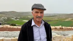 يقف مرزوق أبو نعيم مرتدياً قميصاً أزرق وسترة سوداء وقبعة رمادية مربعة أمام حقول خضراء.