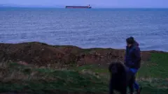 Homem passeia com cachorro em um dia escuro, com um petroleiro no mar ao fundo