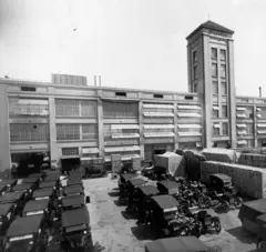 A imagem em preto e branco mostra uma fábrica da Ford vista do lado de fora, com um amplo pátio repleto de automóveis estacionados e caixas de carga. O edifício industrial tem grandes janelas, vários pavimentos e uma torre com o nome da empresa em destaque. A cena retrata uma instalação de produção e distribuição de veículos nas primeiras décadas do século 20.