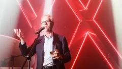 Дерк Сауэр в костюме (без галстука) с бокалом в руке стоит перед микрофоном и жестикулирует. За ним — светящихся символы XX на красном фоне.