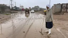 رجل مسن يقف في شارع غمرته مياه الفياضانات والطمي ويرتدي جلباباً وحذاءً بلاستيكياً أبيض اللون يغطي أسفل ركبتيه.