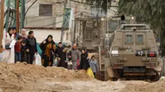 فلسطينيون يسيرون بجوار مدرعات إسرائيلية أثناء مغادرتهم مخيم جنين في الضفة الغربية التي تحتلها إسرائيل، في 22 يناير/كانون الثاني 2025