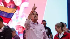Venezuela Devlet Başkanı Nicolás Maduro, yakalı bir gömleğin kollarını sıyırmış ve altına beyaz bir tişört giymiş halde, dev bir ekranın önünde durarak gökyüzünü işaret ediyor.
