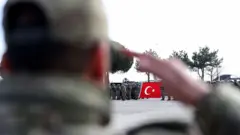 Arkası dönük bir askerin selam yaptığı kolunun arasından Türk bayrağı tutan tabur uzakta görülüyor.
