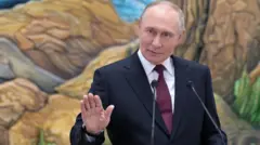 Путін у темному піджаку та бордовій краватці стоїть перед двома мікрофонами та жестикулює рукою