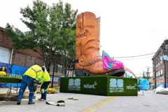 Una calle en Houston, Texas en la que se está instalando una zona para que fanáticos del fútbol puedan disfrutar los partidos. Hay ds trabajadores instalando unas mesas y detrás de ellos se ve una inmensa bota texana de adorno que está instalada en la mitad de la calle