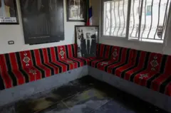 No salão de hóspedes da família Al-Radwan, com bancos de concreto cobertos por tecido preto e vermelho com motivos árabes, a bandeira drusa ao centro e retratos antigos nas paredes.