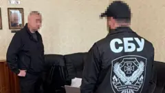 Помещение со столом и черным кожаным диваном вдоль стены.
В кадре двое людей: мужчина с размытым лицом стоит у стола, другой — спиной к камере, в куртке с большой надписью «СБУ» и эмблемой на спине. На столе виден ноутбук с размытым логотипом.
