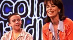 Jane Kaczmarek, who has brown shoulder length hair and is wearing an orange cardigan, sitting next to Frankie Muniz, who is wearing an orange T-shirt underneath a white and brown checked shirt, at a press event for Malcolm in the Middle.