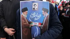 عکسی از تجمعی در حمایت از مجتبی خامنه‌ای؛پوستری در دست یکی از طرفداران حکومت طراحی شده که در آن علی خامنه‌ای (چپ)، رهبر پیشین، در حال تحویل دادن پرچم تا شده ایران به پسرش مجتبی خامنه‌ای (راست) دیده می‌شود.