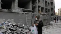 Umugore wambaye agapfukamunwa n'ikanzu y'umukara, witandiye mu mutwe, nyuma y'ibitero bya Israel mu burengerazuba bwa Irani. Mu kiganza kimwe afite telefone igendanwa naho mu kindi kiganza afite ishashi y'umweru n'ubururu irimo ibintu imbere. 