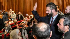 Suriye Devlet Başkanı Ahmed Şara, Ekim ayında başkent Şam'daki bir oy kullanma merkezini ziyaret ediyor. Ve telefonlarıyla fotoğrafını çeken bir grup gazeteciye el sallıyor. Takım elbise ve kravat giymiş ve etrafı diğer erkeklerle çevrili.