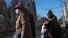 बिजिंगच्या रस्त्यावर मास्क घालून उभी असलेली एक वृद्ध महिला