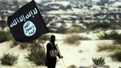 Un militante enmascarado del grupo Estado Islámico posa sosteniendo la bandera de la IE en algún lugar de los desiertos de Irak o Siria en 2015.