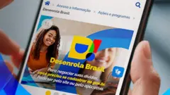 Mãos segurando um celular com logotipo do programa Desenrola Brasil na tela