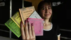 Uma mesária exibe cédulas de votação em verde, amarelo, laranja, azul e rosa. Ela veste uma camiseta preta