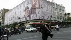 Tahran'daki bir ana caddede Hamaney posteri olan bir binanın önünden geçen bir kadın ve motosiklet üzerinde insanlar ve diğer araçlar görülüyor