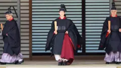 Принц Хисахито в черном облачении для взрослых членов семьи стоит между двумя мужчинами в черном.