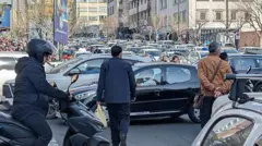 Iranianos tentam atravessar rua em meio ao tráfego intenso em Teerã, capital do Irã, no dia 28 de fevereiro de 2026, após ataques de Israel e EUA. Um homem está em uma scooter e dois outros a pé ao lado de filas de carros.