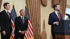 マイクの載った演台に手を添えて話すスーツ姿のヴァンス氏を、後ろからウィトコフ氏とクシュナー氏が見ている。星条旗が並んでいる。