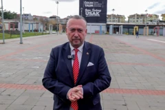 Özkan Yalım kent meydanında ayakta duruyor, ellerini birbirine bağlamış, kameraya bakıyor. 