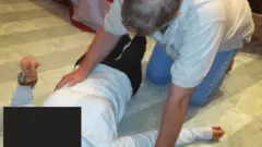 Um homem ajoelhado sobre uma mulher deitada no chão. O rosto dela está oculto por um retângulo preto. Ela veste calças pretas e uma blusa branca e levantou o braço esquerdo. O homem está tocando a lateral do abdômen dela.