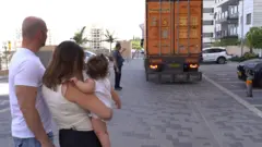 Um caminhão de mudanças laranja dá ré em uma rua enquanto Nofar e Eyal observam sua aproximação. O casal é visto de costas — ele veste uma camiseta branca, e ela, uma blusa bege sem mangas, segurando a filha pequena no colo.